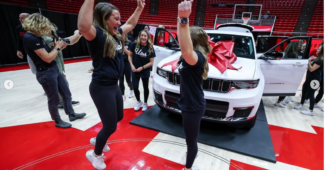 Utah athletes celebrate around a white vehicle with a red bow on it.